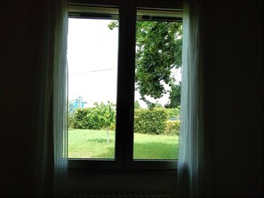 Janela com cortinas leves, vista para um gramado verde e uma árvore.