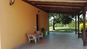 Uma área de varanda coberta com duas cadeiras de vime, uma pequena mesa, um vaso de planta e piso de azulejos.