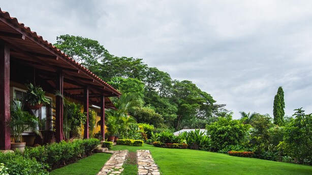 A stone pathway leading to a building with a red roof and columns, surrounded by lush greenery and a well-maintained lawn.