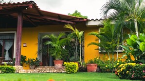 A yellow house with a stone foundation, a red roof, and a garden with various plants and flowers.