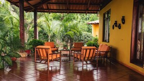 A covered patio with a tiled floor, surrounded by lush greenery, featuring a yellow wall and wooden furniture.