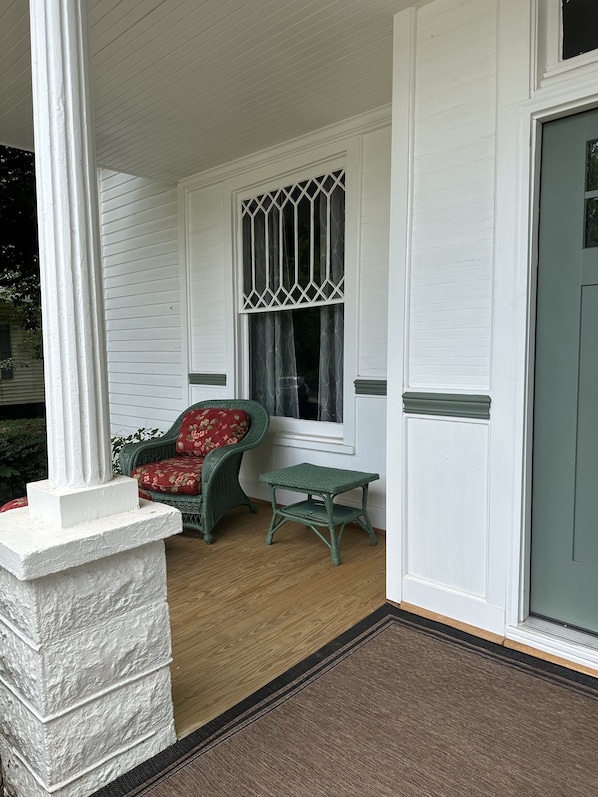 A porch with a wicker chair and a small table, a white door, and a window with a decorative grille.