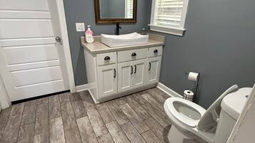 Un baño con lavamanos blanco, espejo, inodoro y ventana con persianas.