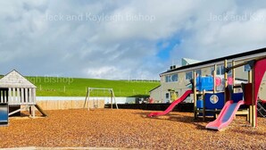 Ein Spielplatz mit einem hölzernen Spielhäuschen und Rutschen, umgeben von einem Holzzaun und einem grasbewachsenen Hügel im Hintergrund.