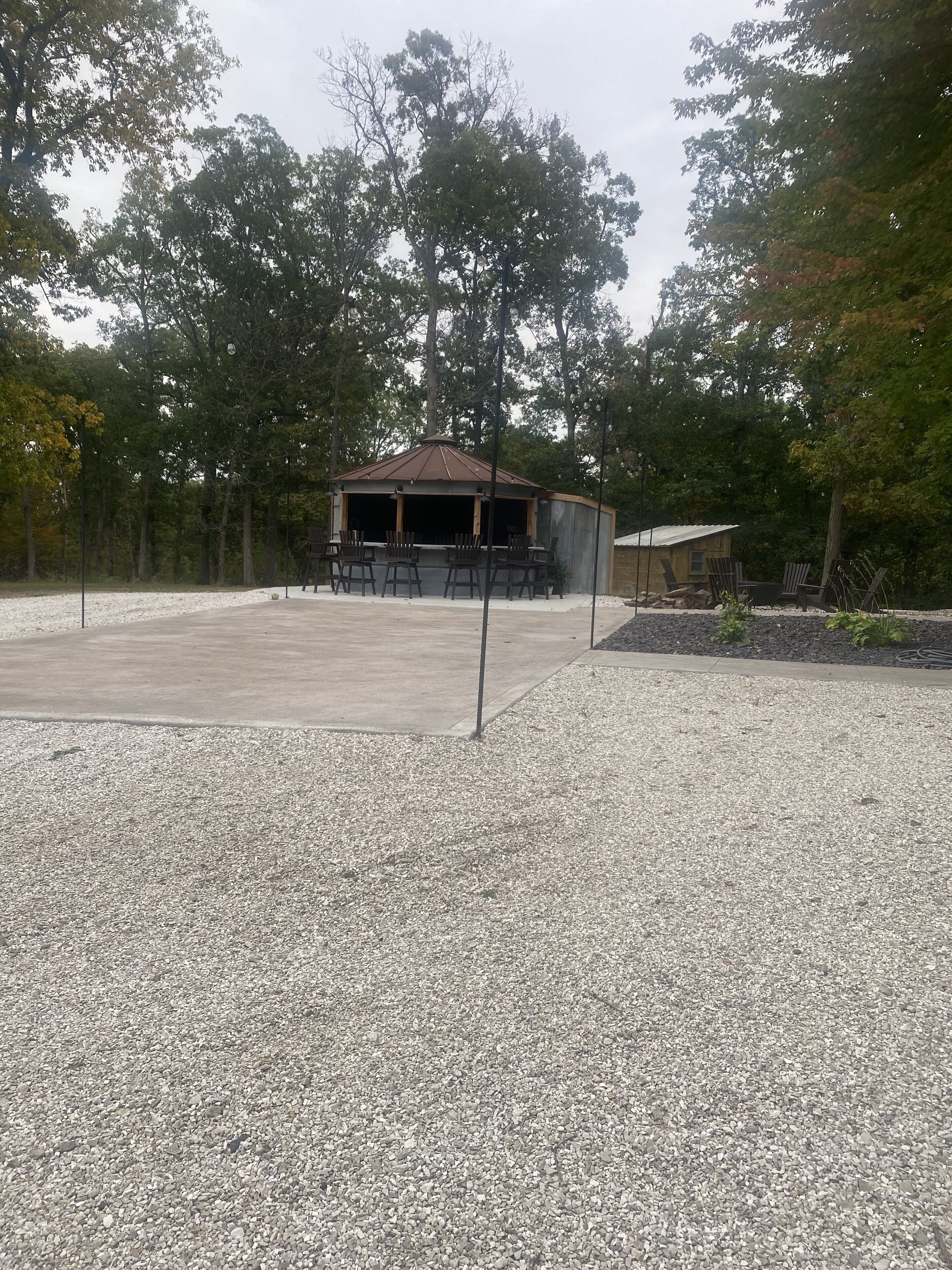 Un gazebo con tetto in metallo e sedie di legno, immerso tra ghiaia e alberi.