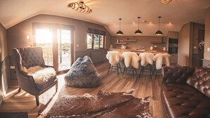 A spacious loft-style living area with a large window, a dining table with chairs, a leather sofa, and a fluffy pouf. - Zen Jungle Retreat (Holsworthy)
