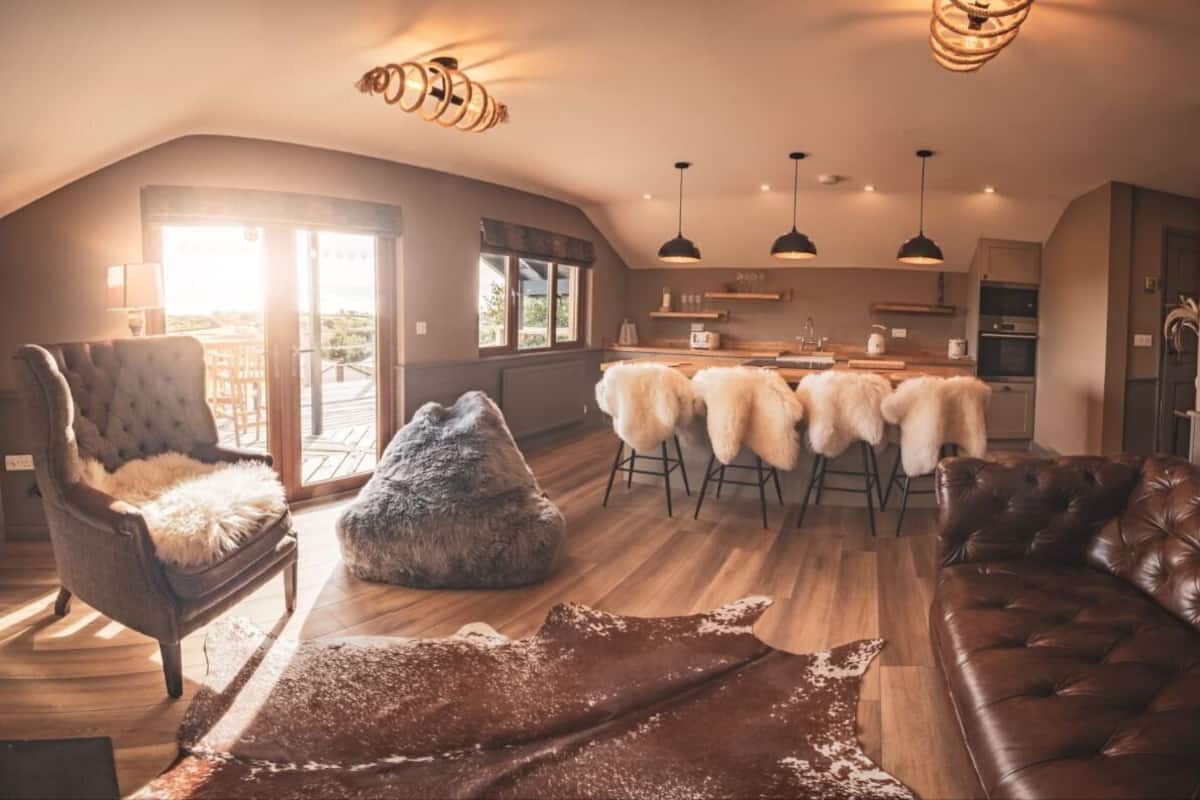 A spacious loft-style living area with a large window, a dining table with chairs, a leather sofa, and a fluffy pouf.
