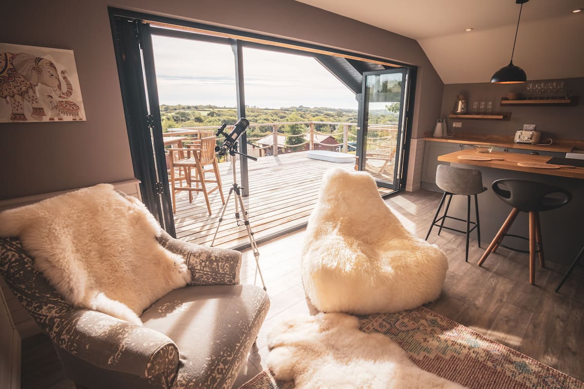 A modern living room with a large sliding glass door leading to an outdoor deck, a cozy seating area with a fluffy white pouf, and a kitchen area with bar stools and a hanging pendant light.