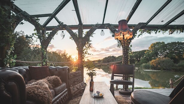 A cozy outdoor seating area with a wooden table, leather sofa, and chairs, overlooking a calm body of water and a sunset. - Zen Jungle Retreat (Holsworthy)
