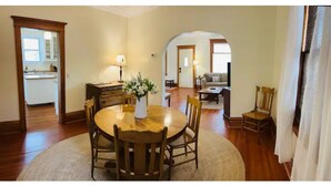 Un comedor con una mesa redonda de madera, sillas y un jarrón con flores. Se ve una cocina al fondo, a través de un arco.