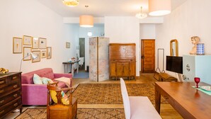 Ein Wohnzimmer mit einem rosa Sofa, einem Holzkabinett und einem Fernseher. An der Wand befinden sich gerahmte Bilder und auf dem Boden ein Teppich.