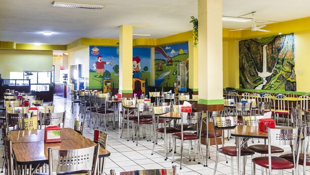 Uma cafeteria com várias mesas e cadeiras, um ventilador de teto e murais coloridos nas paredes.