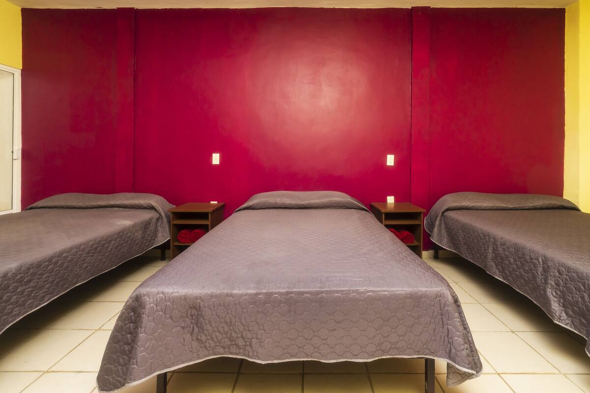 A room with three beds, each with a grey quilted cover, against a red wall. There are two bedside tables with lamps.