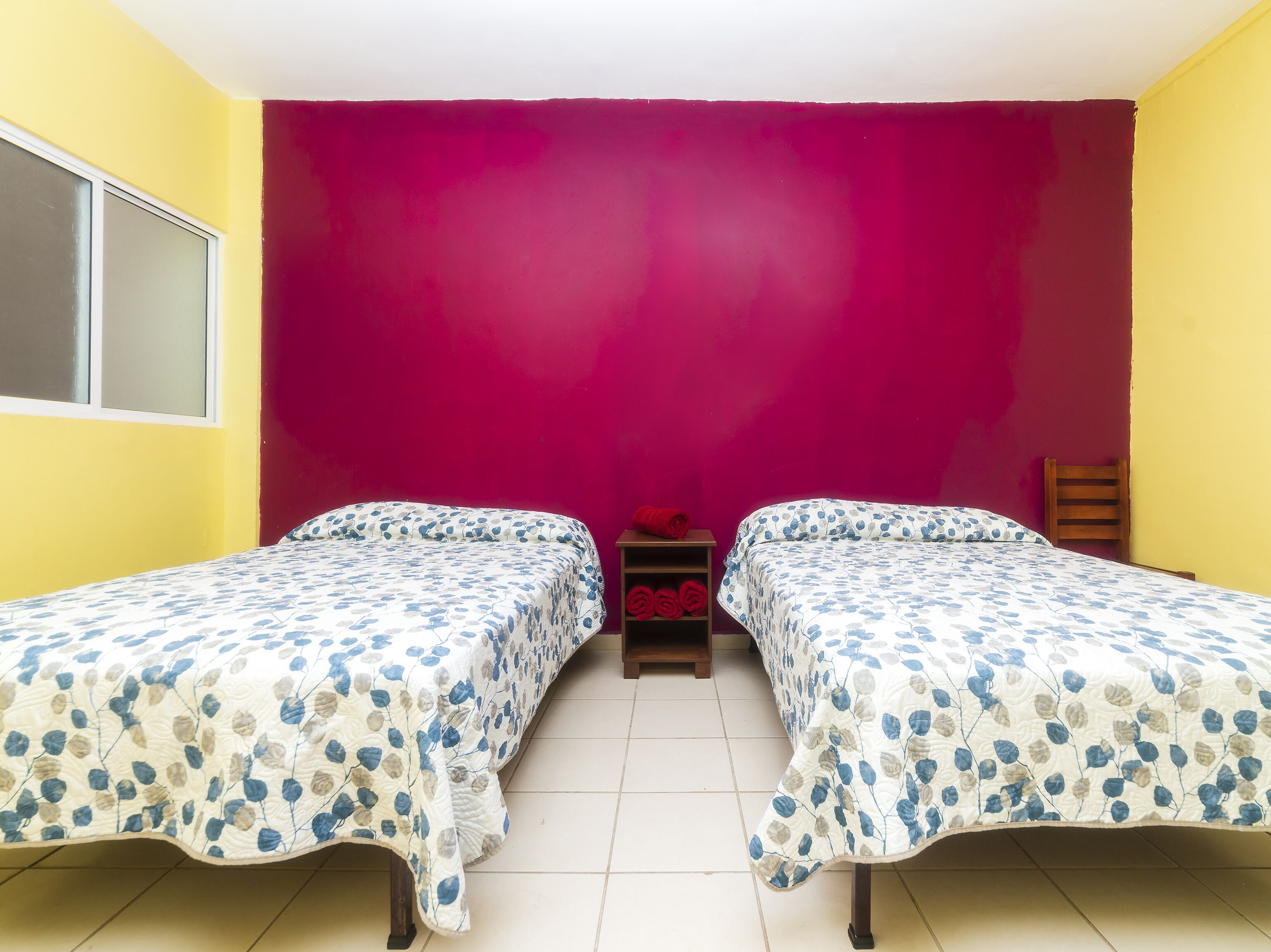 Una stanza con due letti, ognuno con una coperta a motivi floreali, un comodino di legno con un oggetto rosso e una finestra con vetro smerigliato.