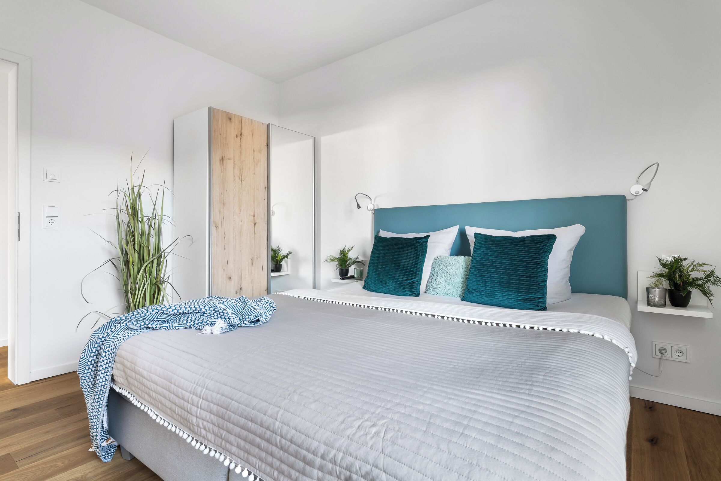 A modern bedroom with a large bed, a wooden wardrobe, and a small shelf with plants.