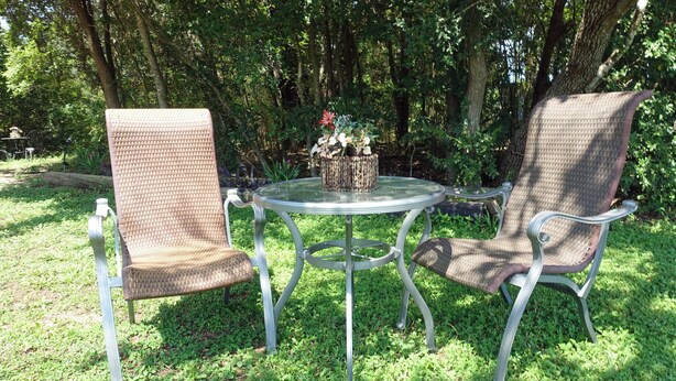 Un área de estar al aire libre con dos sillas de mimbre y una mesa de vidrio, rodeada de follaje.