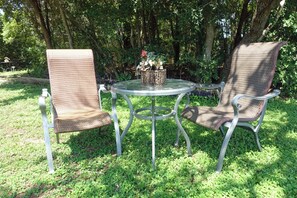 An outdoor seating area with two wicker chairs and a glass table, surrounded by greenery.