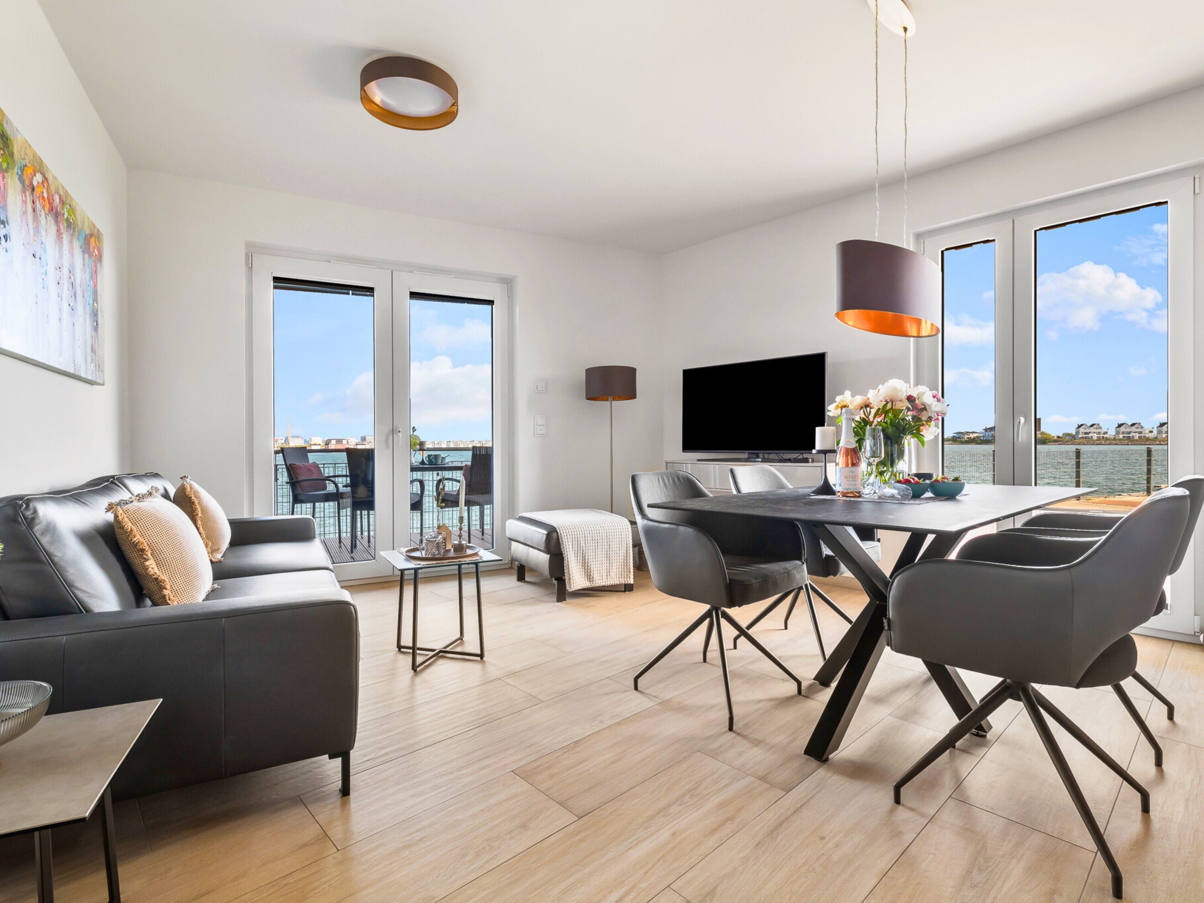 Un salon moderne avec un canapé, une table à manger et des chaises. On y trouve également un téléviseur et une grande fenêtre donnant sur l’eau.