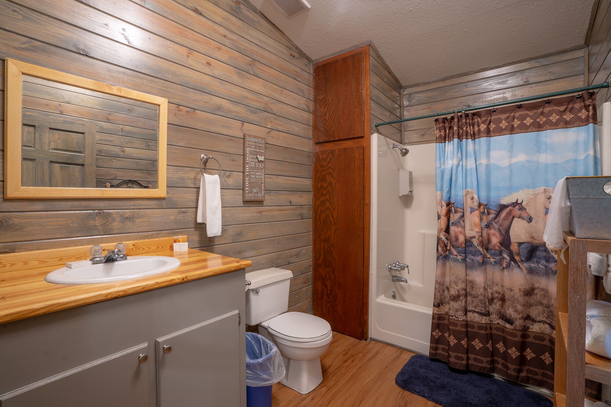 Une salle de bain avec un lambris en bois, un lavabo, des toilettes et un rideau de douche orné d’un motif équestre.