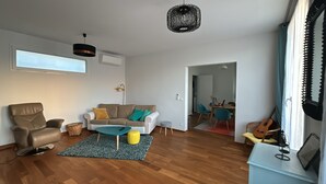 Una sala de estar moderna con un sofá, un sillón y una mesa de centro. Se observa un comedor con sillas y una mesa. Una guitarra es visible a la derecha.