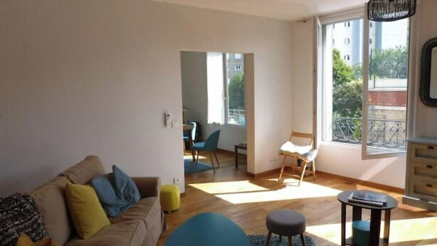Una sala de estar moderna con un sofá, un reposapiés y una mesita. Hay un balcón con barandilla y vistas a edificios y árboles.