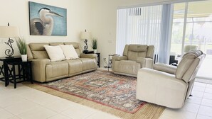A spacious living room with a beige sofa, a white armchair, and a patterned rug. There is a painting of a bird on the wall, a lamp on a side table, and a large window with blinds. - Luxury Retreat/Heated Pool/2Kings /n.Beach/Office (Rotonda West)