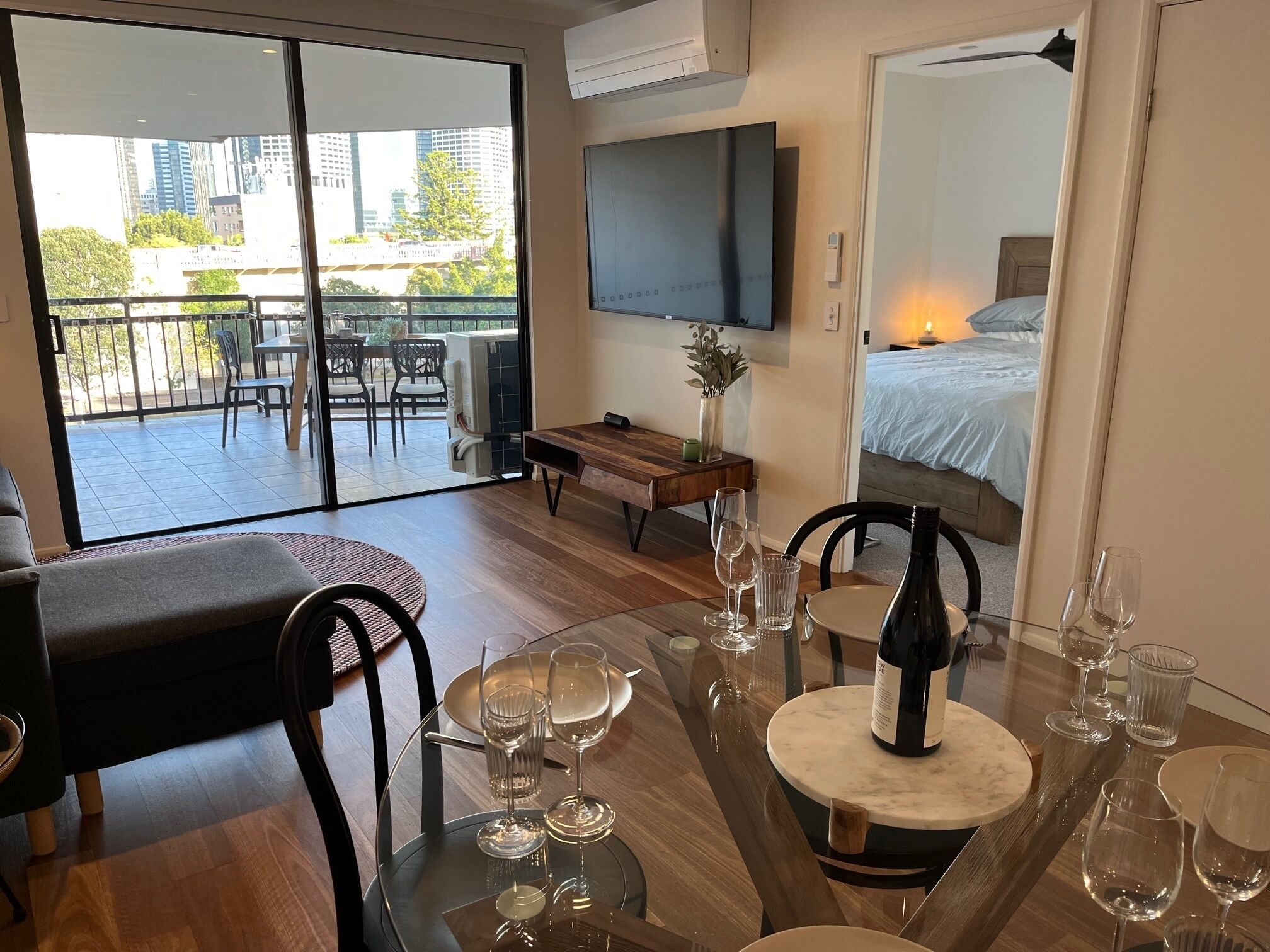 A modern hotel room with a balcony, a dining area with a table set for two, and a bed with a nightstand.