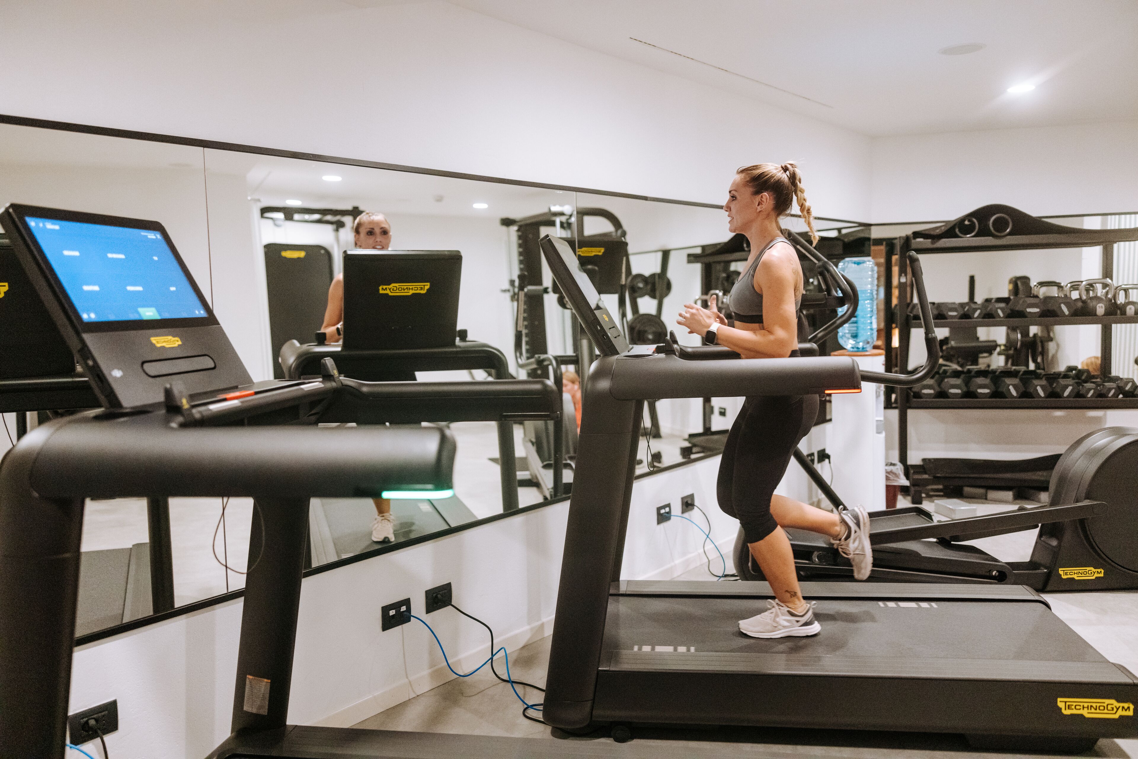 Une femme court sur un tapis de course dans une salle de sport, avec des appareils d’exercice et un écran numérique en arrière-plan.