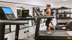 Une femme court sur un tapis de course dans une salle de sport, avec des appareils d’exercice et un écran numérique en arrière-plan.