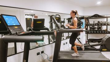 Une femme court sur un tapis de course dans une salle de sport, avec des appareils d’exercice et un écran numérique en arrière-plan.