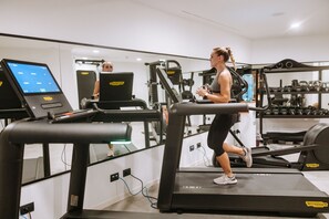 Eine Frau läuft auf einem Laufband in einem Fitnessstudio mit Trainingsgeräten und einem digitalen Bildschirm im Hintergrund.