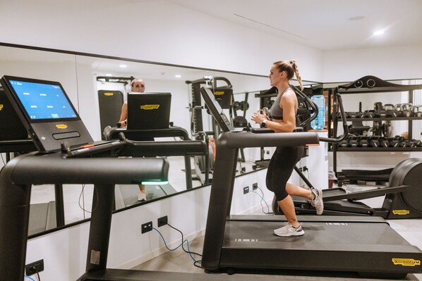 Eine Frau läuft auf einem Laufband in einem Fitnessstudio mit Trainingsgeräten und einem digitalen Bildschirm im Hintergrund.