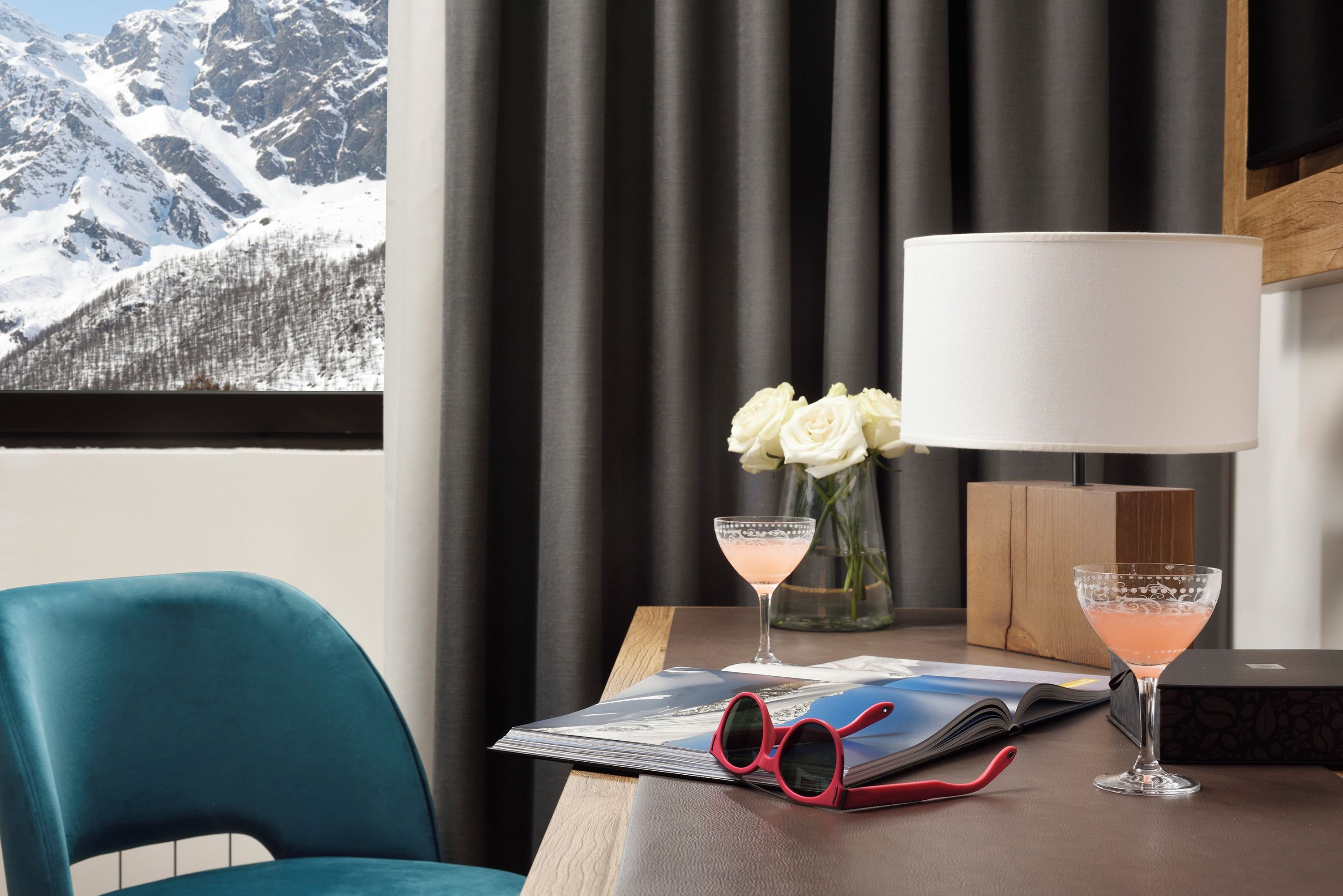 Una stanza con vista su montagne innevate, un tavolo con una rivista, un paio di occhiali da sole rossi, un vaso con fiori bianchi e due bicchieri da cocktail.
