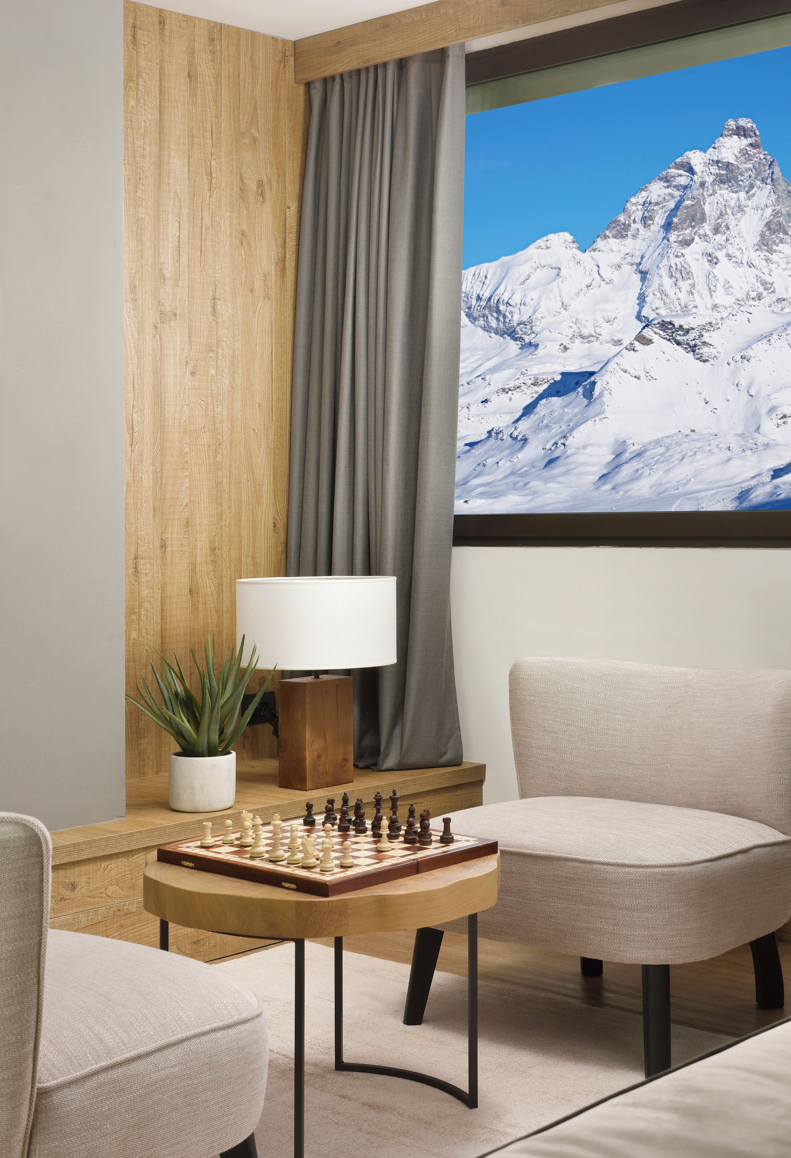 A modern hotel room with a wooden accent wall, a large window with a mountain view, a chess table, and two upholstered chairs.