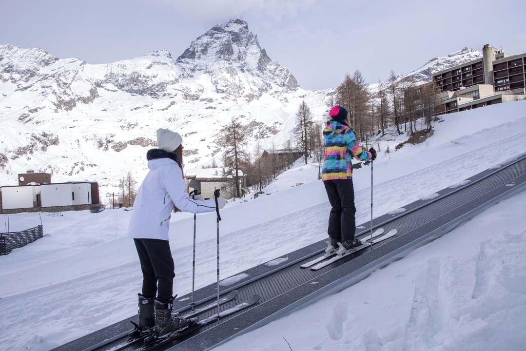 Two skiers on a ski lift with a snow-covered mountain in the background.