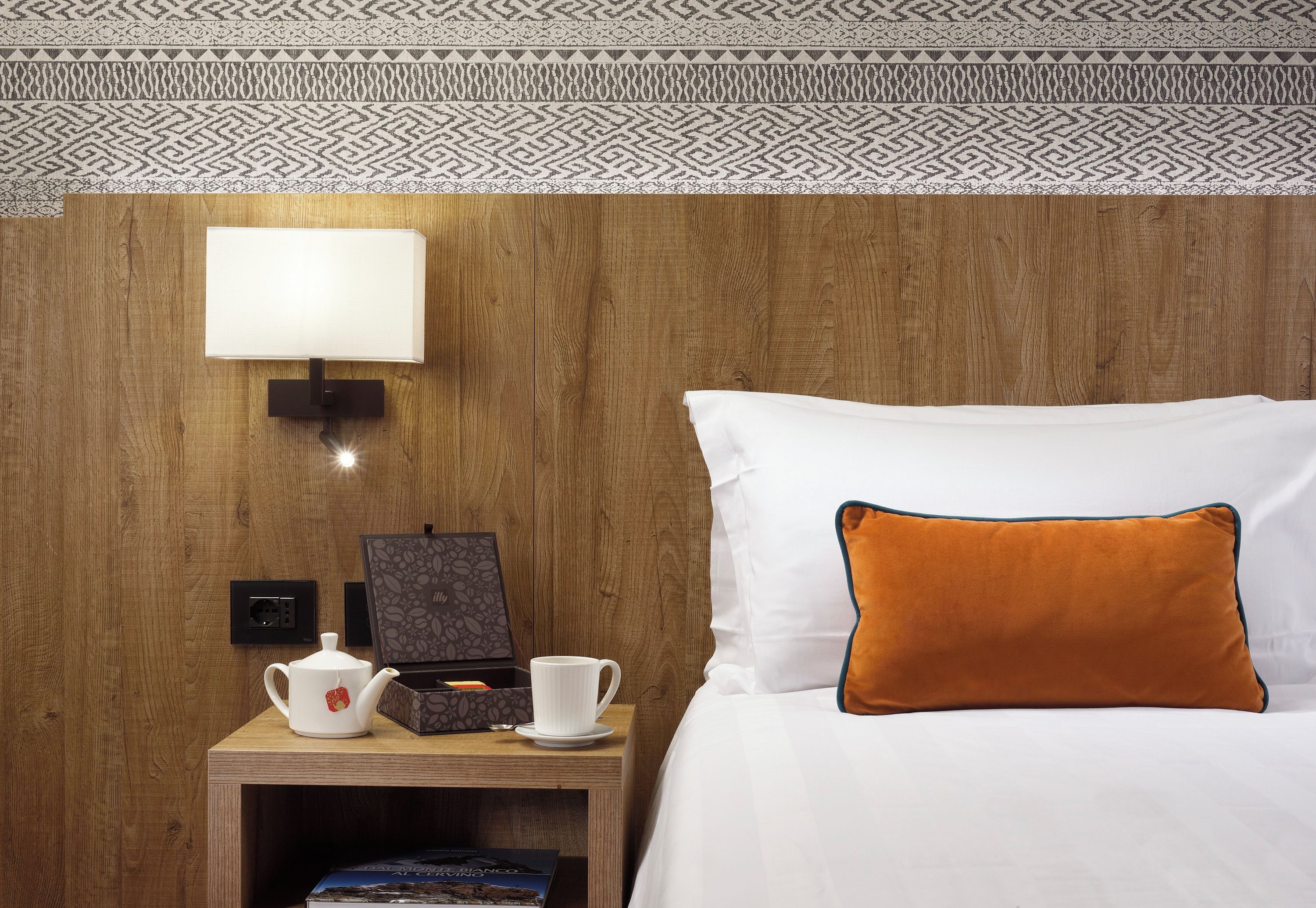 A hotel room with a wooden headboard, a white bed with an orange pillow, a bedside table with a white teapot, a cup, and a book.