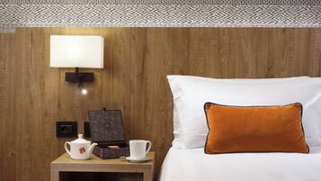 A hotel room with a wooden headboard, a white bed with an orange pillow, a bedside table with a white teapot, a cup, and a book.