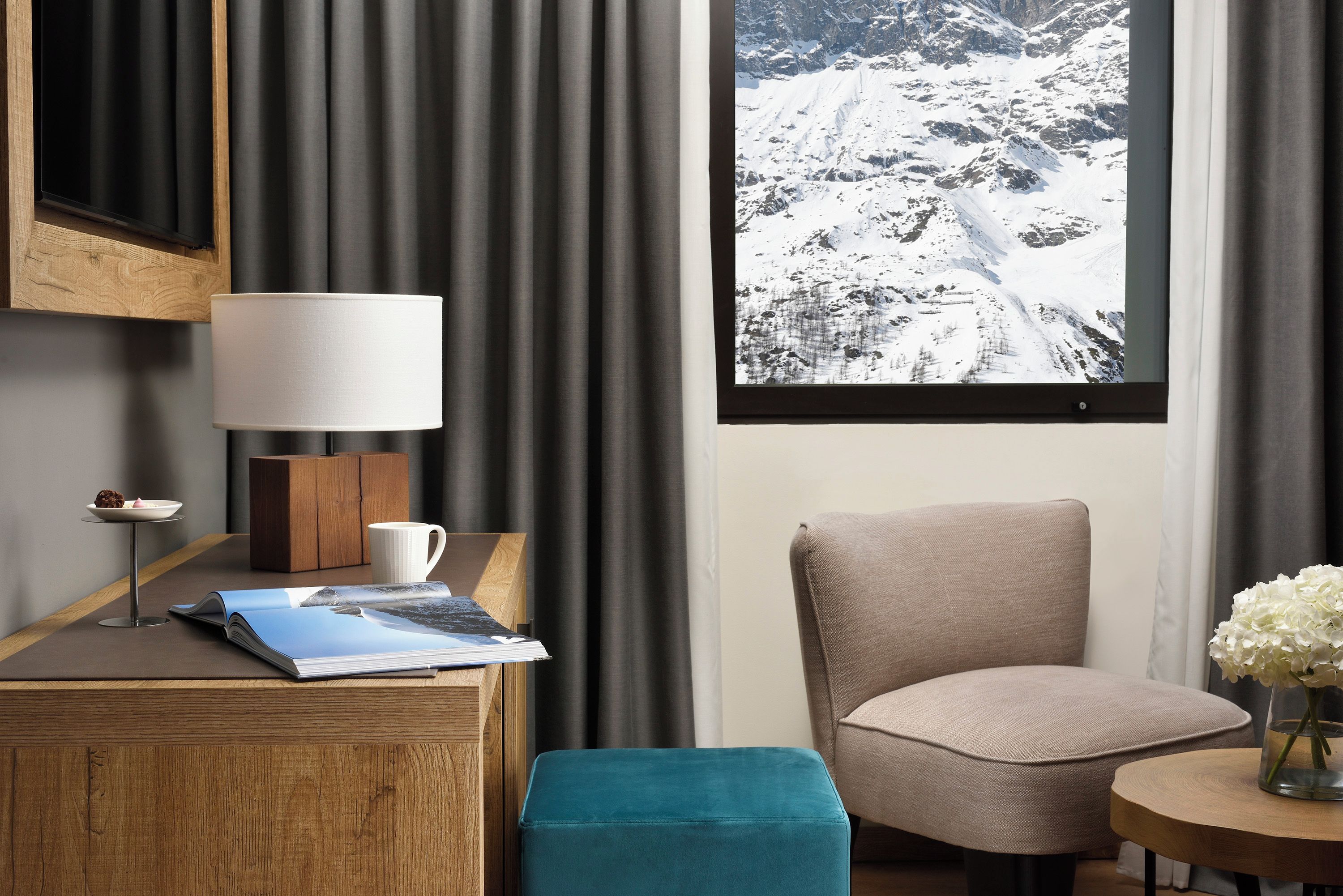Une chambre d’hôtel avec un bureau en bois, une lampe, une tasse blanche, un magazine, un pouf turquoise, un fauteuil beige, une petite table ronde avec un vase de fleurs et une fenêtre donnant sur une montagne enneigée.