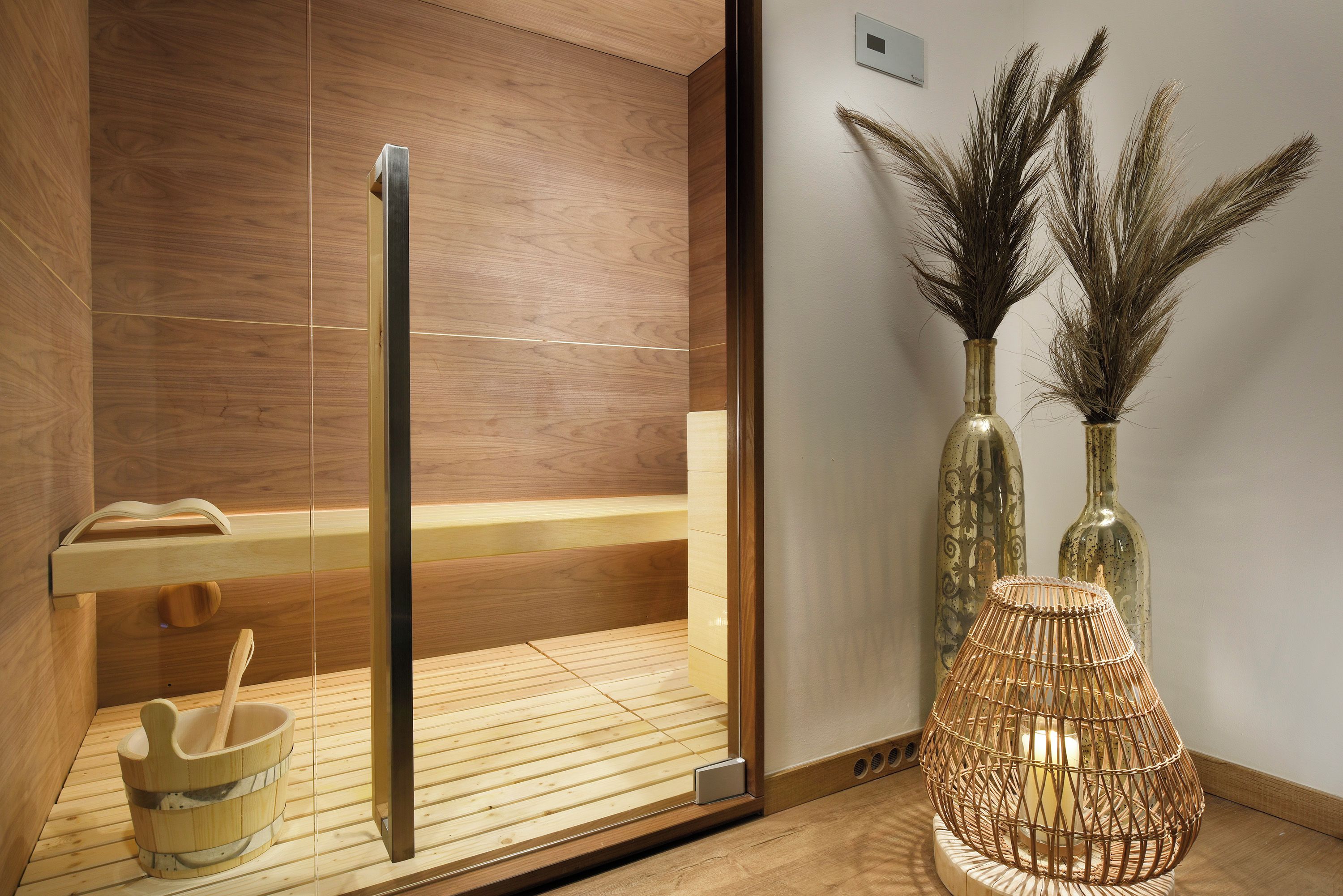 A sauna room with wooden flooring, a glass door, a wooden bench, a bucket, and two decorative vases with dried plants.