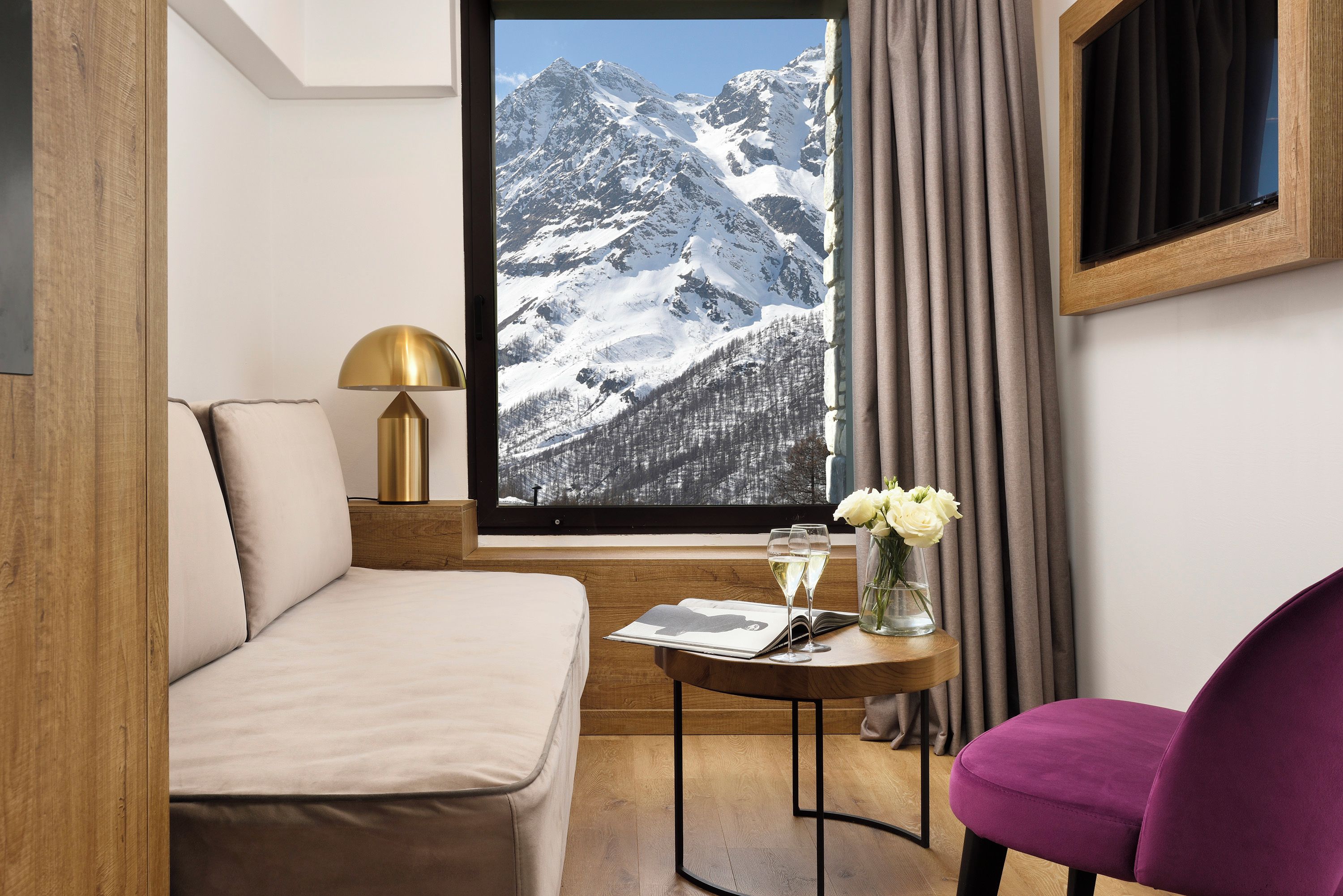 A hotel room with a bed, a window with a mountain view, a table with a vase of flowers, and a purple chair.