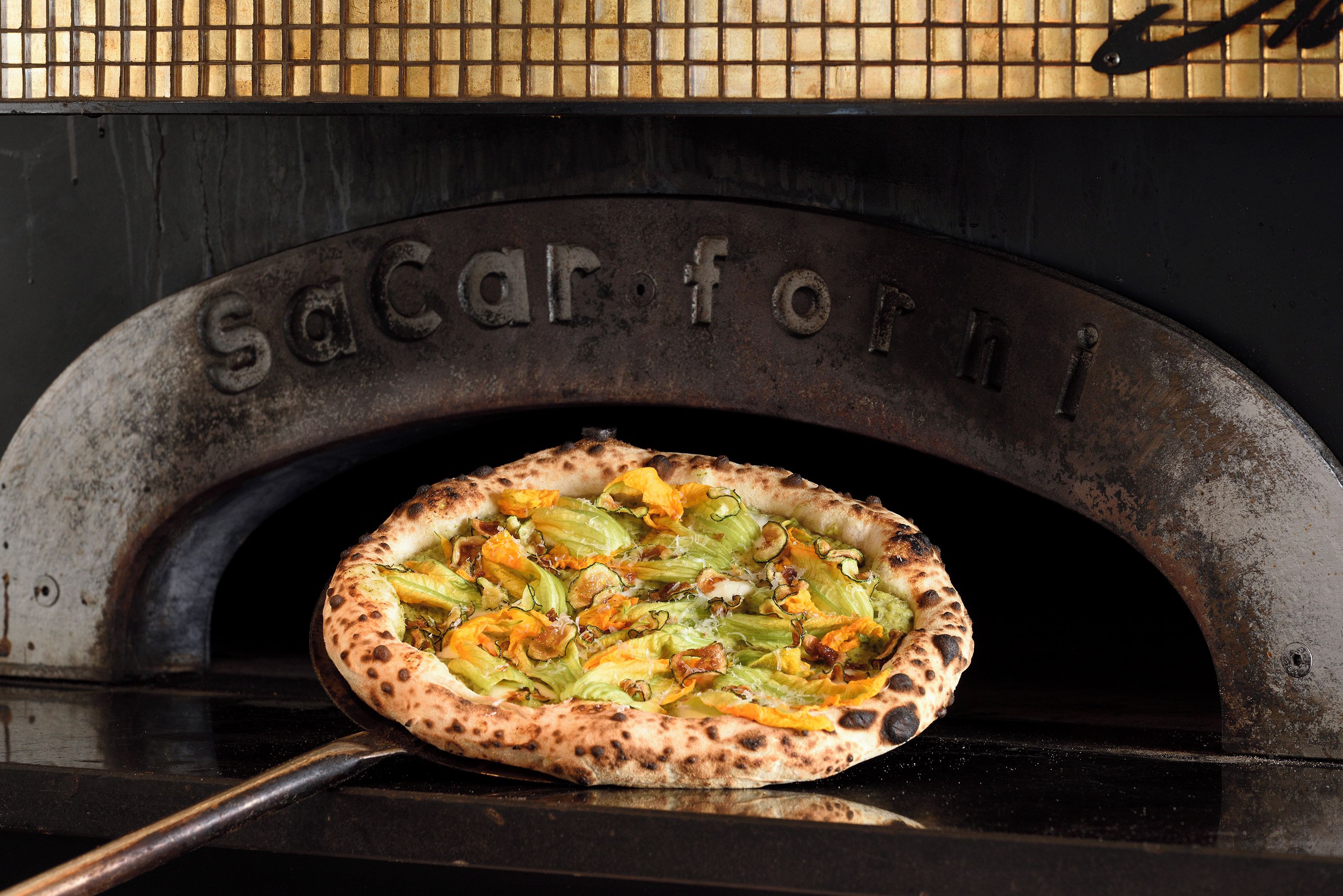 Une pizza cuite dans un four en briques, sur la porte duquel on peut lire l’inscription « Sacarforni ».