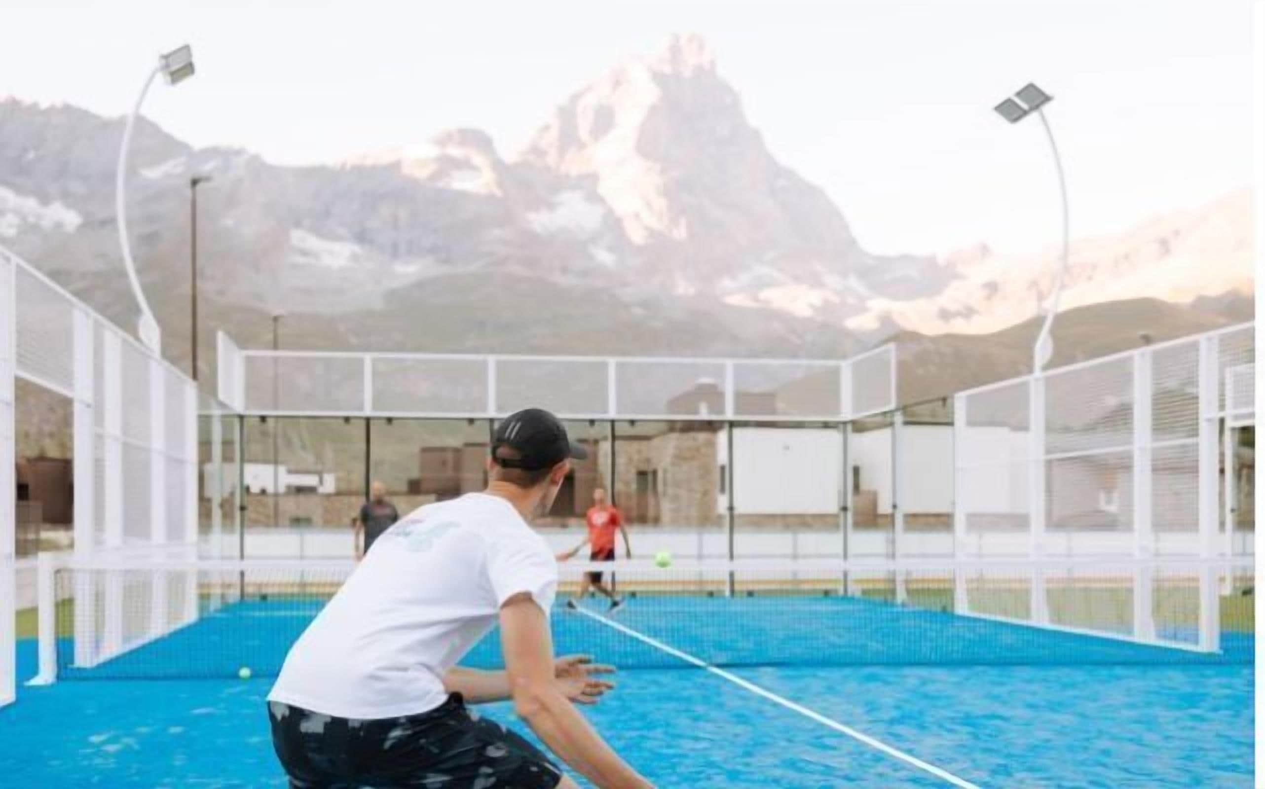 Instalación deportiva interior con una cancha de tenis cubierta por una red, rodeada de paredes de vidrio y con montañas al fondo.