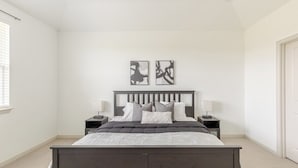 Een slaapkamer met een donker houten bed, twee nachtkastjes met lampen, een raam met zonwering en twee abstracte kunstwerken aan de muur.