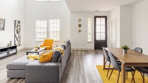 Een ruime woonkamer met een grijze modulaire bank, een gele fauteuil, een houten eettafel met stoelen, en een groot raam dat natuurlijk licht binnenlaat.