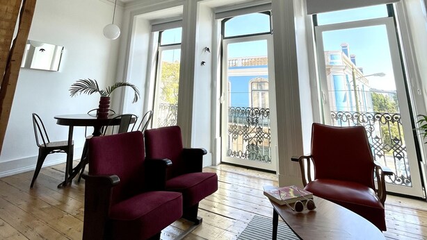 A spacious living room with a wooden floor, a red sofa, a small table, and a balcony with a view of buildings.