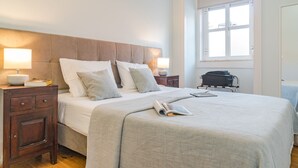 A neatly made bed with a grey bedspread and pillows, a bedside table with a lamp, and a suitcase on a chair.