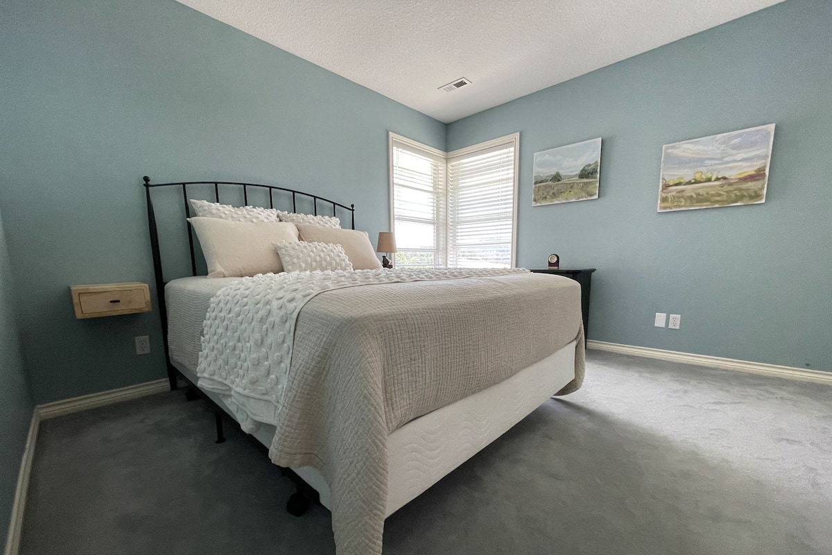 A bedroom with a bed, a nightstand, a window with blinds, and two wall-mounted pictures.