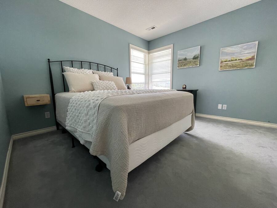 A bedroom with a bed, a nightstand, a window with blinds, and two wall-mounted pictures.