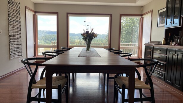 A dining room with a long wooden table and chairs, large windows with a view of the outdoors, and a kitchen area with dark wooden cabinets. - The Gaard House (Carlton)
