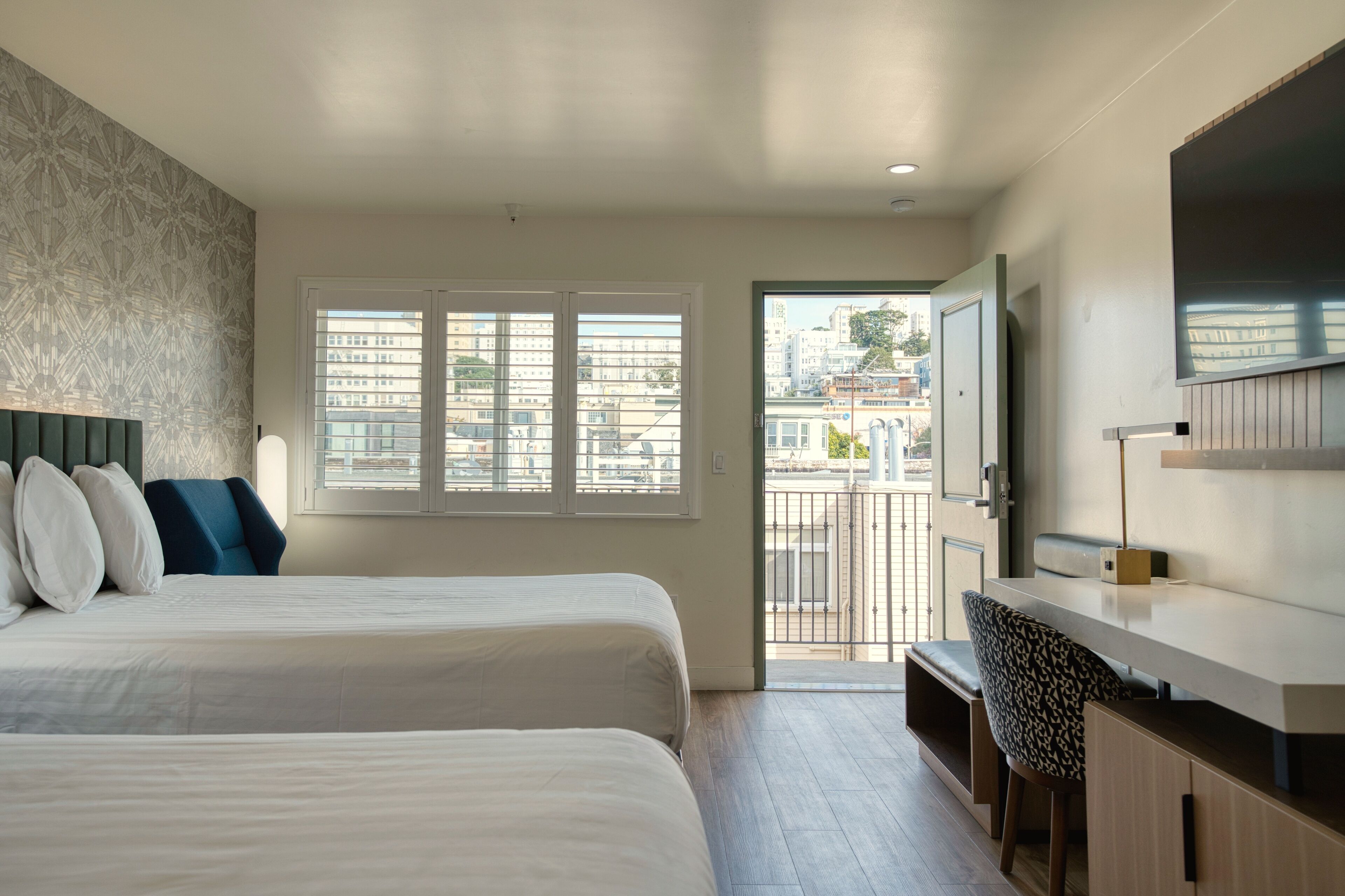Una habitación de hotel moderna con dos camas, un ventanal con persianas, un balcón, un televisor y una zona de escritorio.